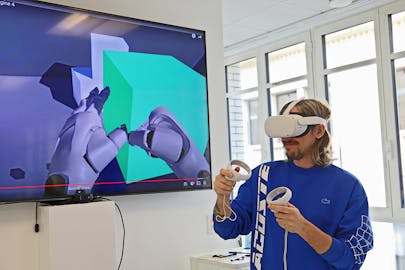 A student wearing a VR headset manipulates a 3D model using two controllers. The view inside the headset is shown on a screen in the background.